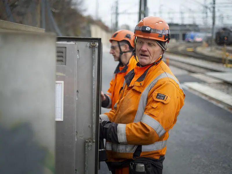 Två stycken banktekniker i orangea skyddskläder byter komponenter i ett elskåp